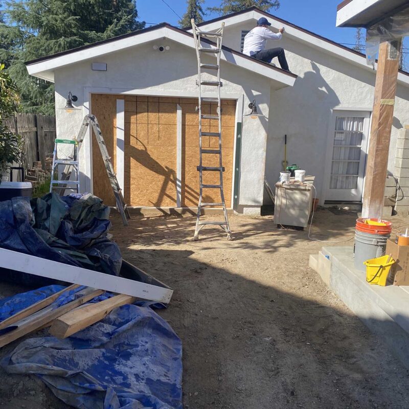 Construction site showing a garage renovation with a worker on the roof and a boarded-up garage door opening.