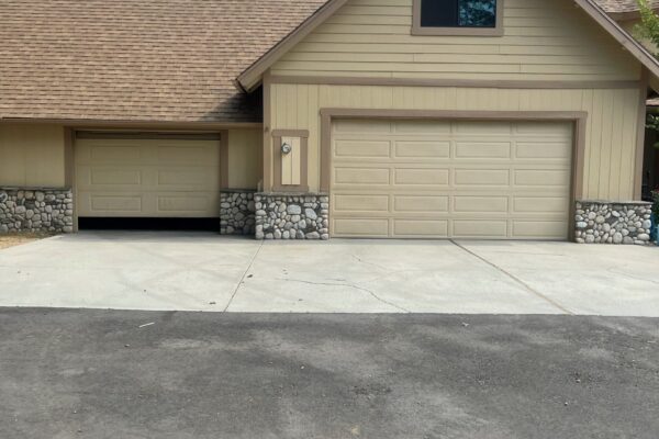 Two tan raised-panel garage doors on a home with beige siding, stone pillar accents, and a wide concrete driveway.