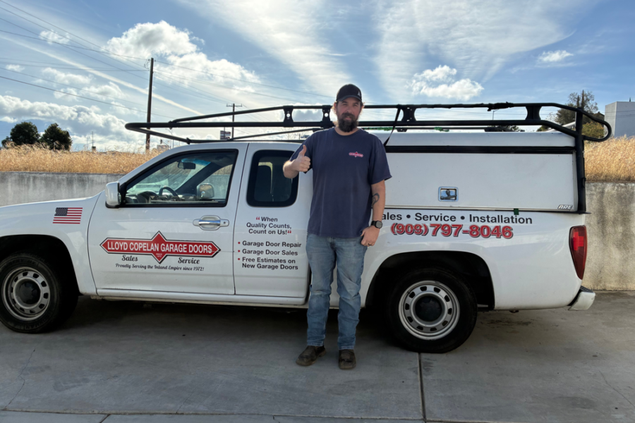 Man in a cap and blue shirt giving a thumbs up beside a white Lloyd Copelan Garage Doors service truck with a roof ladder rack on a concrete lot.